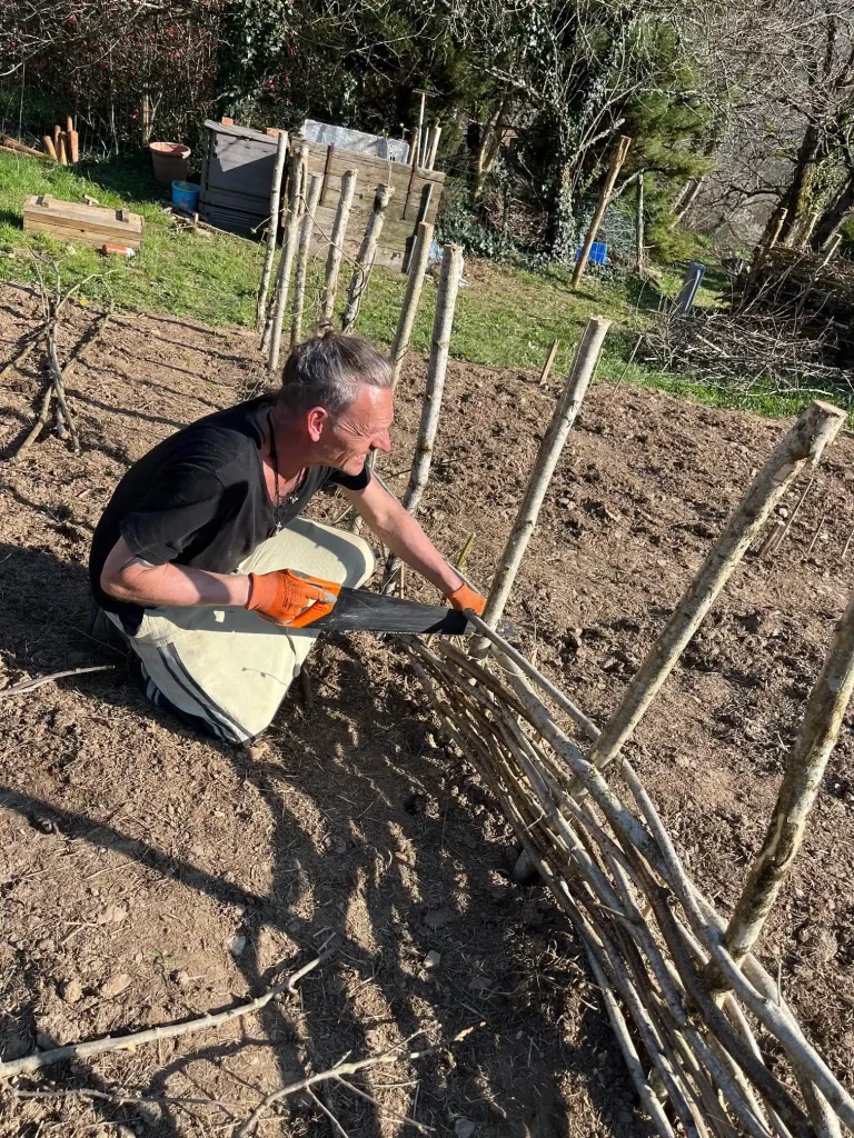 Een man met grijze haren werkt in de moestuin op Domaine du Puech Fleuri, waarbij hij zich buigt over jonge plantaanzettingen met bamboe stokken als ondersteuning. De afbeelding toont praktische landbouwwerkzaamheden die deel uitmaken van de regeneratieve landbouwaanpak van La Nouvelle Terre.