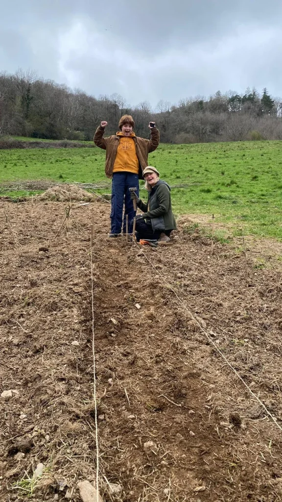 Twee kinderen poseren trots in een moestuin op het biologische domein, waarbij ze groenten planten in de vruchtbare aarde van La Nouvelle Terre in Frankrijk.