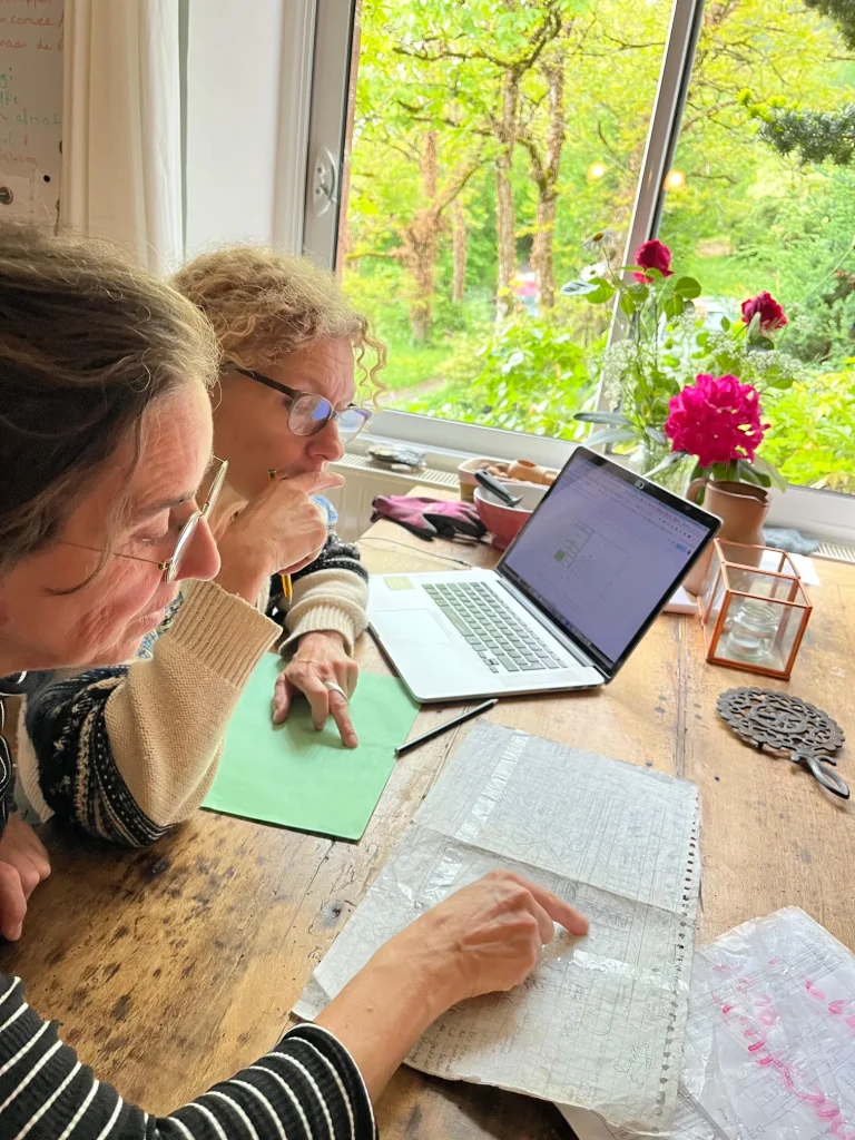 Twee deelgenoten van La Nouvelle Terre werken samen aan planning en administratie bij het raam met uitzicht op het groene domein en de bloeiende rozen.