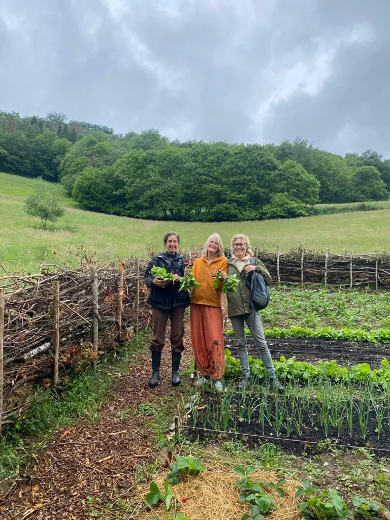 Drie leden van La Nouvelle Terre staan in de moestuin van Domaine du Puech Fleuri met verse groenten, omgeven door permacultuur bedden en houten afrastering met groene weilanden en bos op de achtergrond.